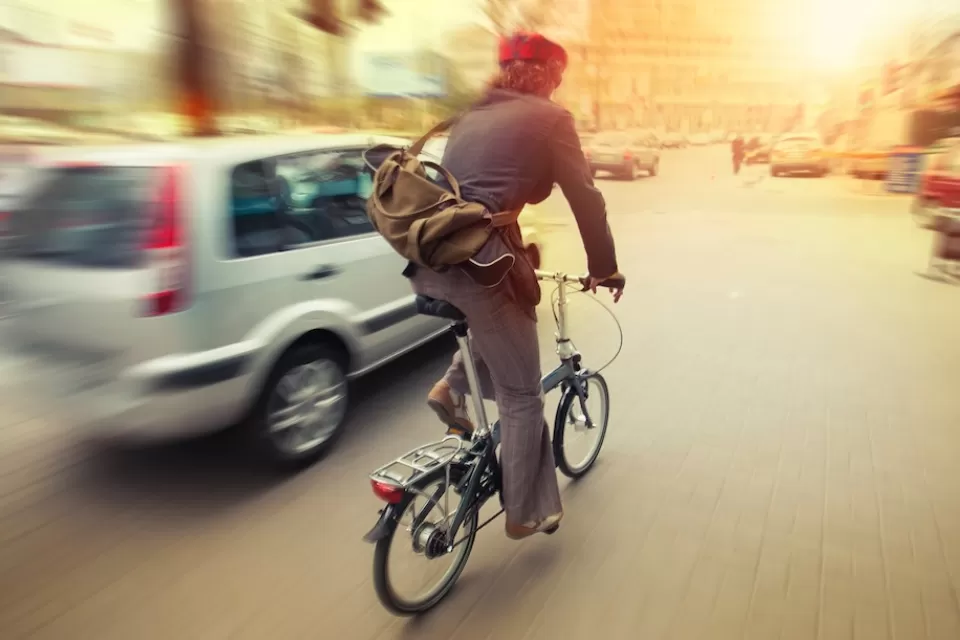 Cyclist on folding bike riding next to silver car on city street.